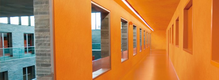 Długi korytarz pokryty pomarańczową farbą znajdujący się w biurowcu i oknami z lewej strony wychodzącymi na dziedziniec.