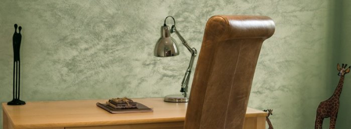 Stylowy gabinet, z dużym skórzanym krzesłem i biurkiem, na którym znajduje się lampka oraz zeszyt. Elementy afrykańskie w postaci figurki na biurku oraz żyrafy w prawym rogu.