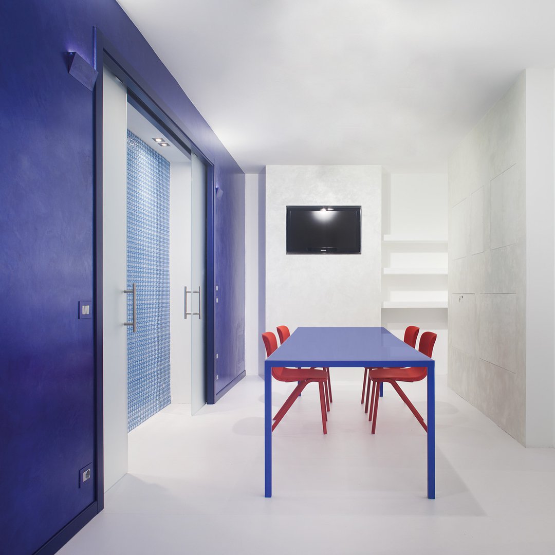 Jadalnia w minimalistycznym stylu o ścianach w kolorze biało-niebieskim. W centralnej części jadalni znajduje się niebieski stół z czerwonymi krzesłami.