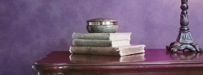 Stare książki leżą na biurku, a obok stoi podstawa od świecznika. Tło wypełnia fioletowa farba.